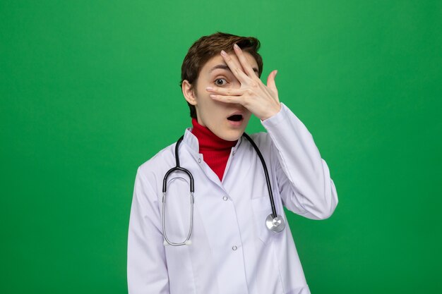 Joven médico en bata blanca con estetoscopio alrededor del cuello sorprendido cubriendo los ojos con los dedos mirando a través de los dedos de pie sobre la pared verde