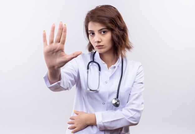 Joven médico en bata blanca con estetoscopio de pie con la mano abierta haciendo señal de stop mirando a la cámara con cara seria sobre fondo blanco.