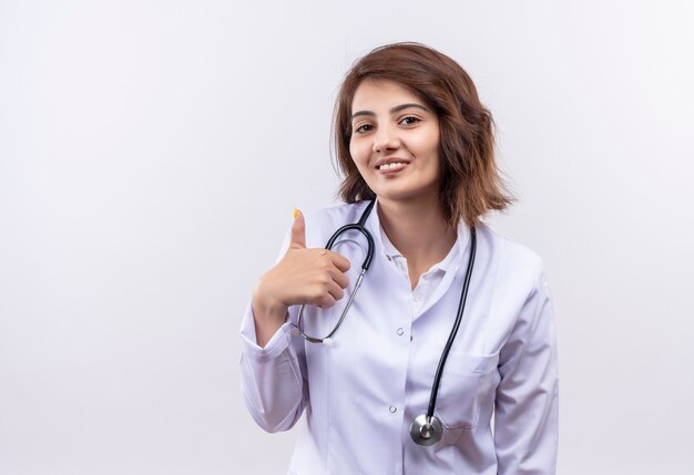 Joven médico en bata blanca con estetoscopio sonriendo mostrando los pulgares para arriba de pie sobre la pared blanca.