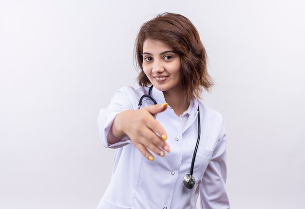 Joven médico en bata blanca con estetoscopio sonriendo saludo confiado ofreciendo una mano de pie sobre la pared blanca