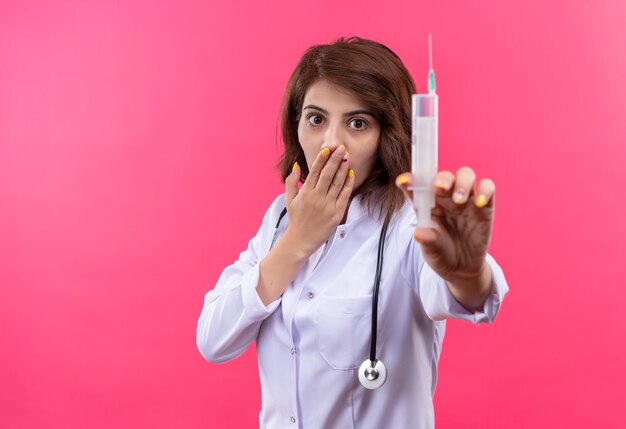 Joven médico en bata blanca con estetoscopio sosteniendo jeringa sorprendido cubriendo la boca con la mano