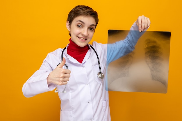 Joven médico en bata blanca con estetoscopio sosteniendo radiografía de los pulmones sonriendo confiado mostrando los pulgares para arriba de pie en naranja