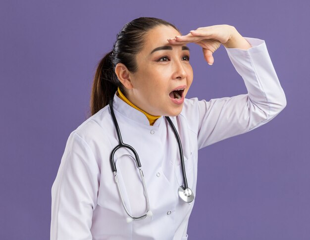 Joven médico en bata blanca de medicina con estetoscopio alrededor del cuello mirando a lo lejos con la mano sobre la cabeza en busca de alguien o algo de pie sobre la pared púrpura