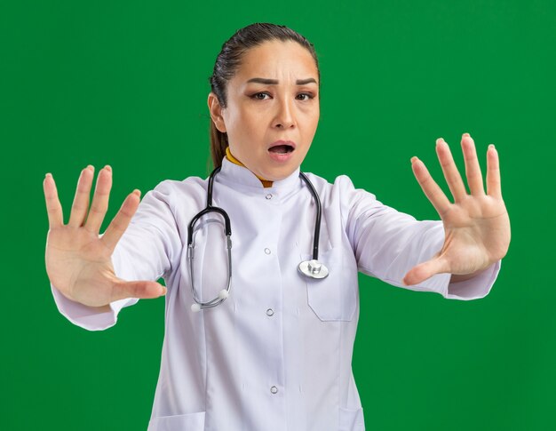 Joven médico en bata blanca de medicina con estetoscopio alrededor del cuello preocupado haciendo gesto de parada con las manos de pie sobre la pared verde