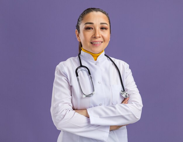 Joven médico en bata blanca de medicina con estetoscopio alrededor del cuello sonriendo confiado con los brazos cruzados de pie sobre la pared púrpura