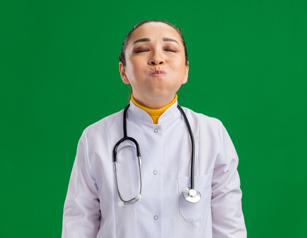 Joven médico en bata blanca de medicina con estetoscopio alrededor del cuello soplando las mejillas con los ojos cerrados de pie sobre la pared verde