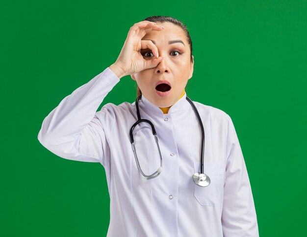 Joven médico en bata blanca de medicina con estetoscopio alrededor del cuello a través de los dedos haciendo el signo de ok estar asombrado de pie sobre la pared verde