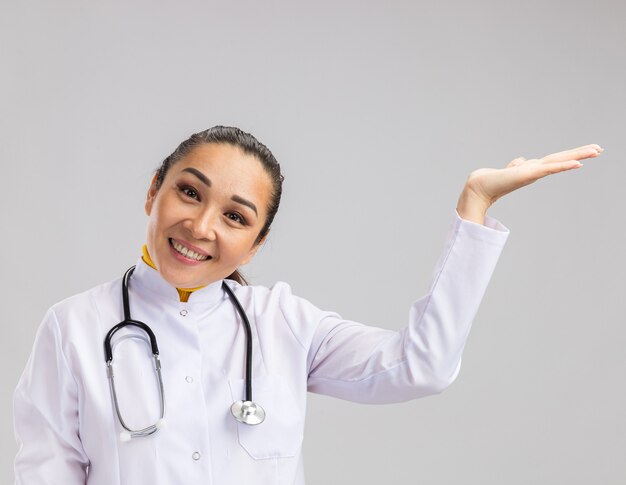 Joven médico en bata médica blanca con estetoscopio alrededor del cuello presentando espacio de copia con brazo sonriendo con cara feliz de pie sobre una pared blanca