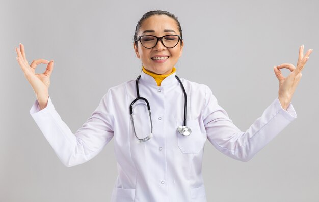 Joven médico en bata médica blanca con estetoscopio alrededor del cuello relajante haciendo gesto de meditación con los dedos de pie sobre la pared blanca