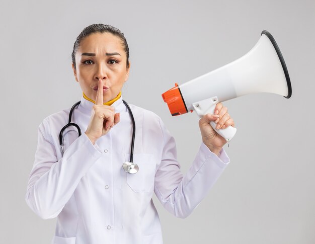 Joven médico en bata médica blanca con estetoscopio alrededor del cuello sosteniendo megáfono haciendo gesto de silencio con el dedo en los labios de pie sobre la pared blanca