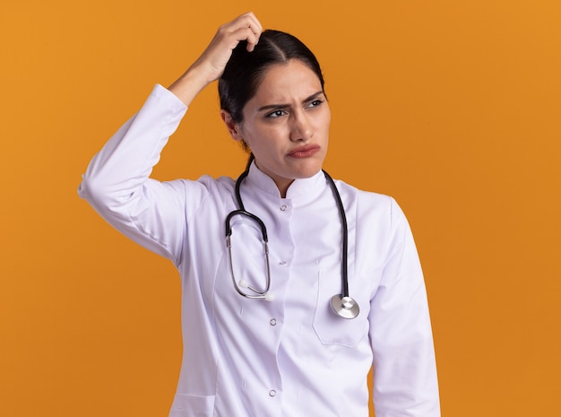 Joven médico en bata médica con estetoscopio alrededor de su cuello mirando a un lado confundido rascándose la cabeza de pie sobre la pared naranja