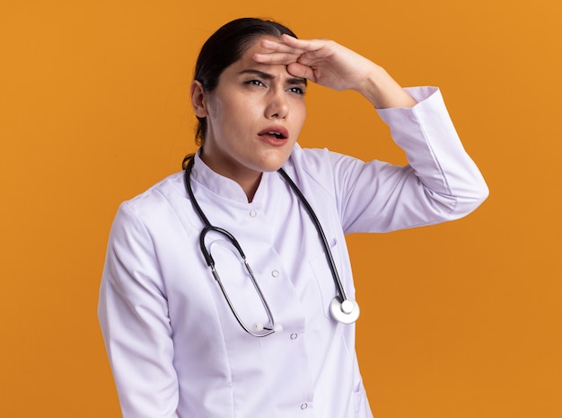 Joven médico en bata médica con estetoscopio alrededor de su cuello mirando a lo lejos con las manos sobre la cabeza de pie sobre la pared naranja