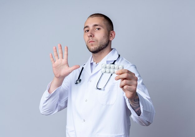 Joven médico con bata médica y un estetoscopio alrededor de su cuello que muestra el paquete de tabletas médicas y cinco con la mano y mirando a cámara aislada sobre fondo blanco