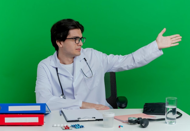 Joven médico con bata médica y estetoscopio con gafas sentado en el escritorio con herramientas médicas girando la cabeza a un lado y apuntando con la mano al lado aislado en la pared verde