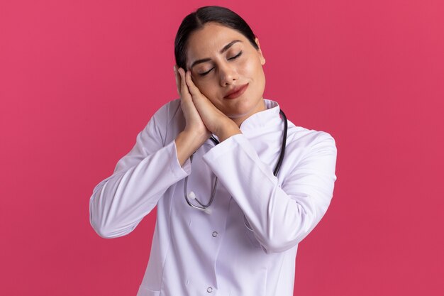 Joven médico en bata médica con estetoscopio haciendo gesto de sueño sosteniendo las palmas juntas inclinando la cabeza en las palmas de pie sobre la pared rosa