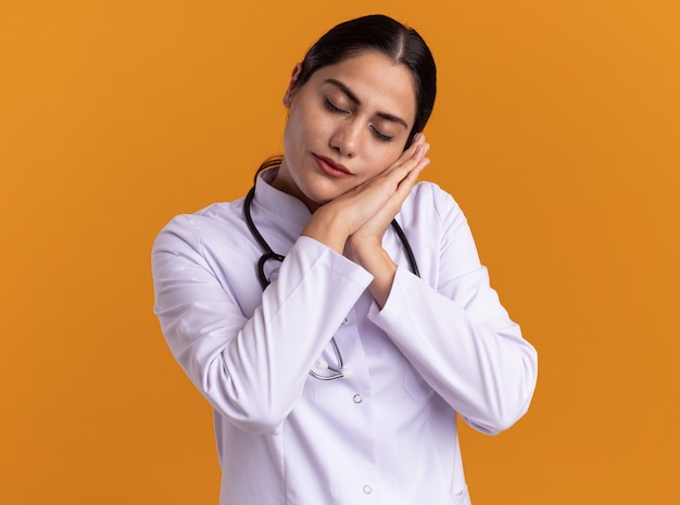 Joven médico en bata médica con estetoscopio haciendo gesto de sueño sosteniendo las palmas juntas inclinando la cabeza sobre las palmas de pie sobre la pared naranja