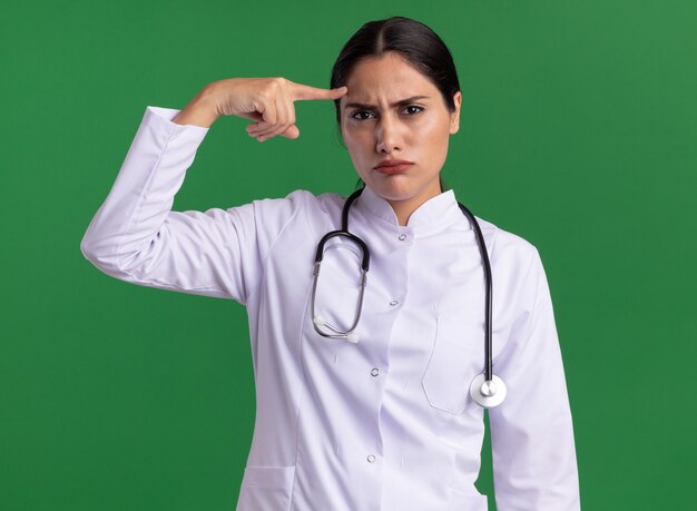 Joven médico en bata médica con estetoscopio mirando al frente con cara seria disgustado señalando con el dedo índice en su sien de pie sobre la pared verde