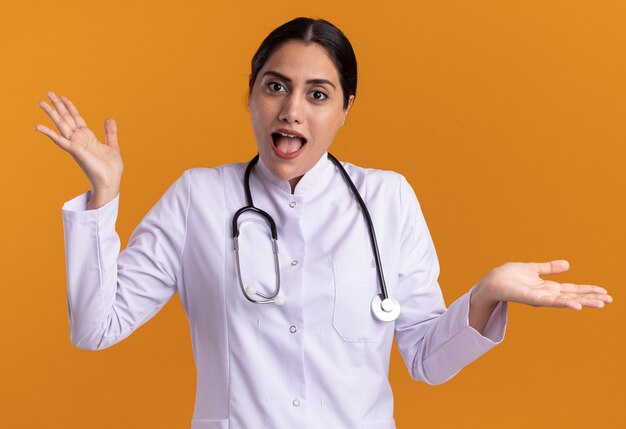 Joven médico en bata médica con estetoscopio mirando al frente confundido extendiendo los brazos a los lados alrededor de su cuello de pie sobre una pared naranja