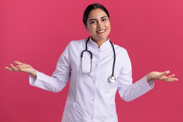 Joven médico en bata médica con estetoscopio mirando al frente sonriendo alegremente con los brazos levantados de pie sobre la pared rosa