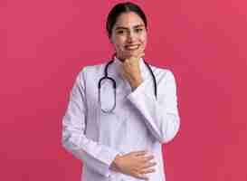 Foto gratuita joven médico en bata médica con estetoscopio mirando al frente sonriendo confiado de pie sobre la pared rosa