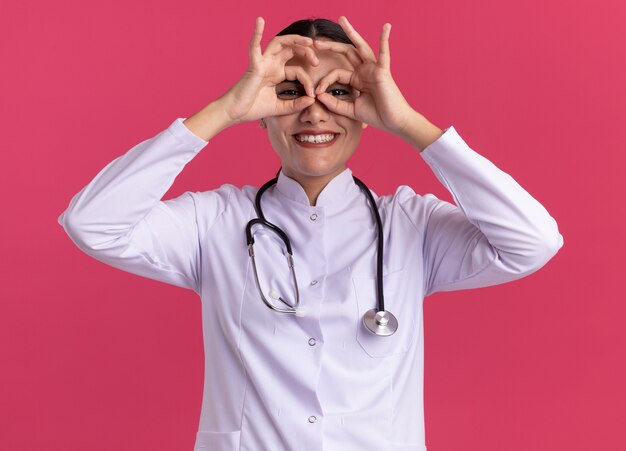 Joven médico en bata médica con estetoscopio mirando al frente a través de los dedos haciendo gesto binocular sonriendo alegremente de pie sobre la pared rosa