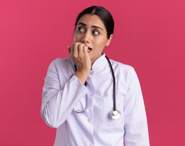 Joven médico en bata médica con estetoscopio mirando a un lado estresado y nervioso morderse las uñas de pie sobre la pared rosa