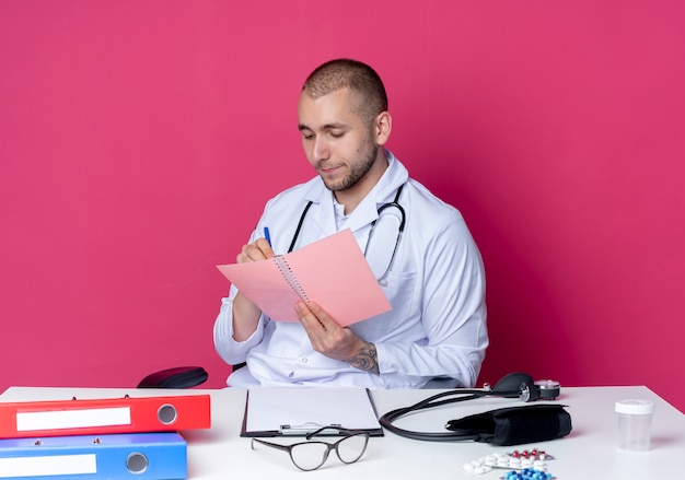 Joven médico con bata médica y estetoscopio sentado en el escritorio con herramientas de trabajo sosteniendo y mirando el bloc de notas y escribiendo algo en él con lápiz aislado sobre fondo rosa