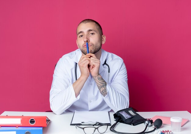 Joven médico con bata médica y un estetoscopio sentado en el escritorio con herramientas de trabajo sosteniendo la pluma y tocando los labios con ella y mirando a cámara aislada sobre fondo rosa