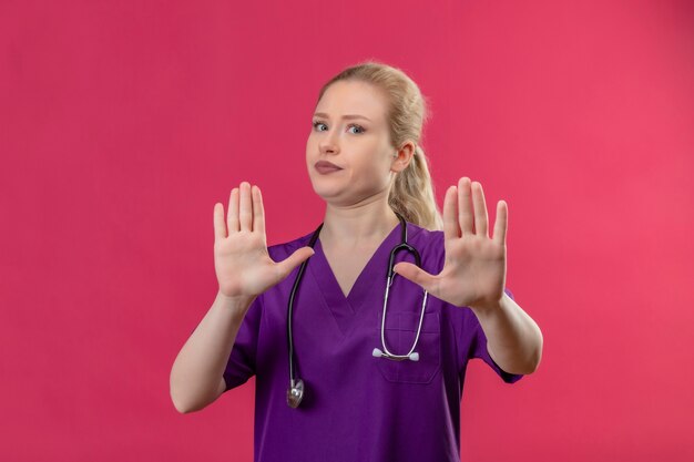Joven médico con bata médica púrpura y estetoscopio muestra gesto de parada en pared rosa aislada
