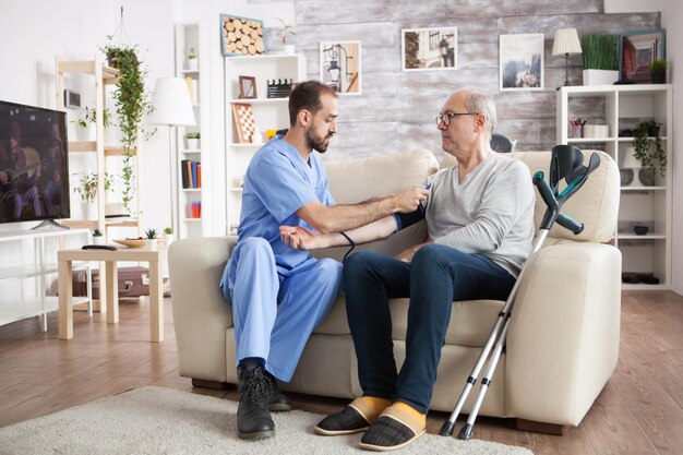 Un joven médico caucásico en un hogar de ancianos revisando la presión arterial de un hombre de edad avanzada con muletas.