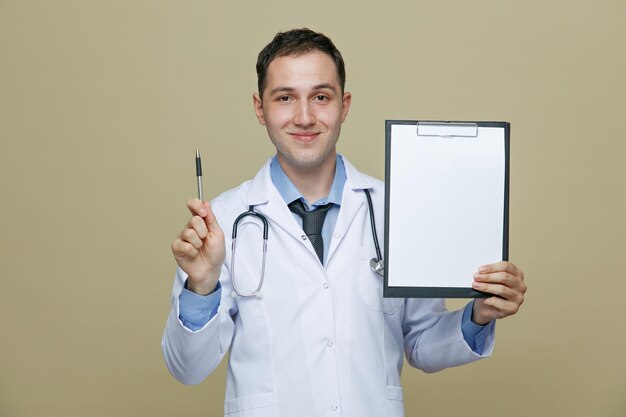 Joven médico complacido con bata médica y estetoscopio alrededor del cuello mirando a la cámara que muestra el portapapeles y la pluma aislados en el fondo verde oliva