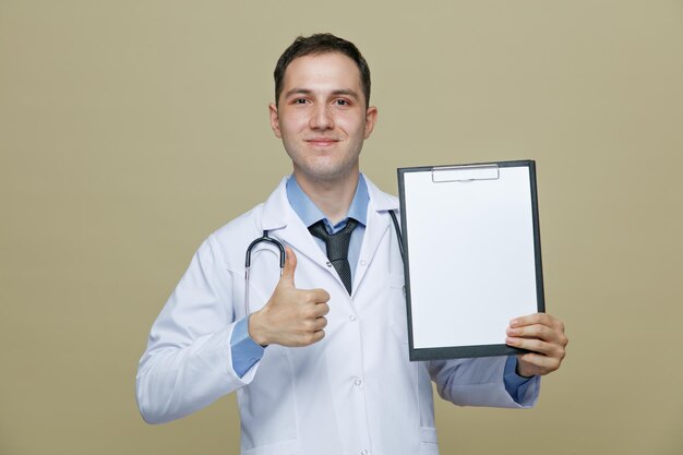 Joven médico complacido con bata médica y estetoscopio alrededor del cuello mirando a la cámara que muestra el portapapeles y el pulgar hacia arriba aislado en el fondo verde oliva