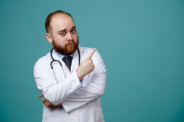 Un joven médico confiado que usa abrigo médico y estetoscopio alrededor de su cuello mirando la cámara apuntando hacia un lado aislado en un fondo azul con espacio para copiar