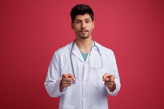 Un joven médico confiado que usa uniforme médico y estetoscopio alrededor de su cuello mirando la cámara que le muestra un gesto aislado en el fondo rojo