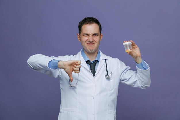 Joven médico descontento con bata médica y estetoscopio alrededor del cuello mirando a la cámara que muestra el recipiente de medida con pastillas y el pulgar hacia abajo aislado en el fondo morado