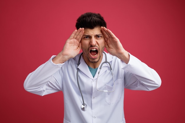 Foto gratuita un joven médico enfadado con uniforme médico y estetoscopio mirando a la cámara con las manos en la cabeza gritando en voz alta algo aislado de fondo rojo