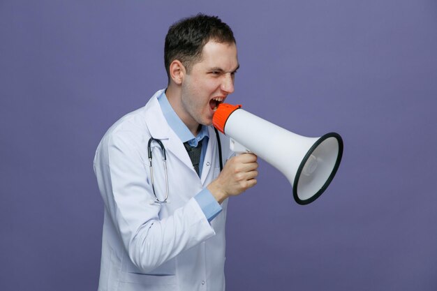 Un joven médico enojado que usa bata médica y estetoscopio alrededor del cuello parado en la vista de perfil mirando al lado hablando en un altavoz aislado en un fondo morado