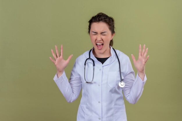 Joven médico enojado vistiendo bata médica con estetoscopio extiende las manos sobre la pared verde