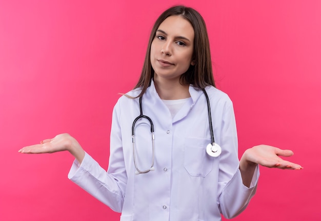 Joven médico con estetoscopio bata médica extiende las manos sobre la pared rosa aislada