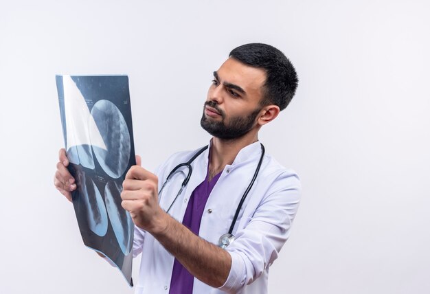 Joven médico con estetoscopio bata médica mirando rayos x en su mano en blanco aislado