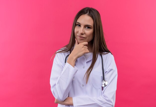 Joven médico con estetoscopio bata médica puso su mano en la barbilla en la pared rosa aislada