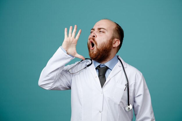 Joven médico frunciendo el ceño con abrigo médico y estetoscopio alrededor del cuello mostrando la mano vacía cerca de la cabeza mirando al costado gritando algo aislado en el fondo azul