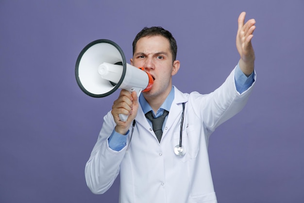 Joven médico frunciendo el ceño con bata médica y estetoscopio alrededor del cuello mirando a la cámara manteniendo la mano en el aire hablando con un locutor aislado en un fondo morado