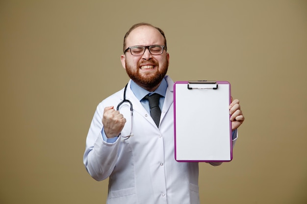 Joven médico frunciendo el ceño con gafas, bata de laboratorio y estetoscopio alrededor del cuello, mostrando el portapapeles y el gesto de sí con los ojos cerrados, aislado en el fondo verde oliva