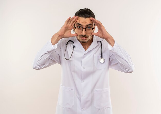 Joven médico con gafas ópticas vistiendo túnica blanca con estetoscopio abriendo los ojos con las manos