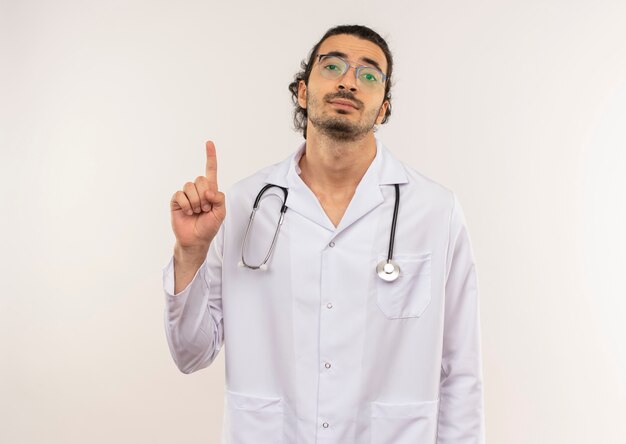 Joven médico con gafas ópticas vistiendo túnica blanca con estetoscopio apunta hacia arriba