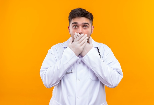 Joven médico guapo con bata médica blanca, guantes médicos blancos y un estetoscopio asustado cerrando su ratón con las manos sobre la pared naranja