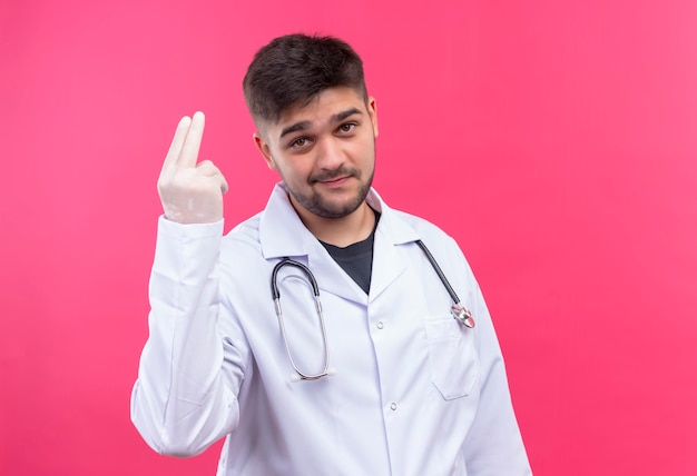 Joven médico guapo con bata médica blanca, guantes médicos blancos y un estetoscopio con cara fría que muestra dos dedos para arriba sobre la pared rosa