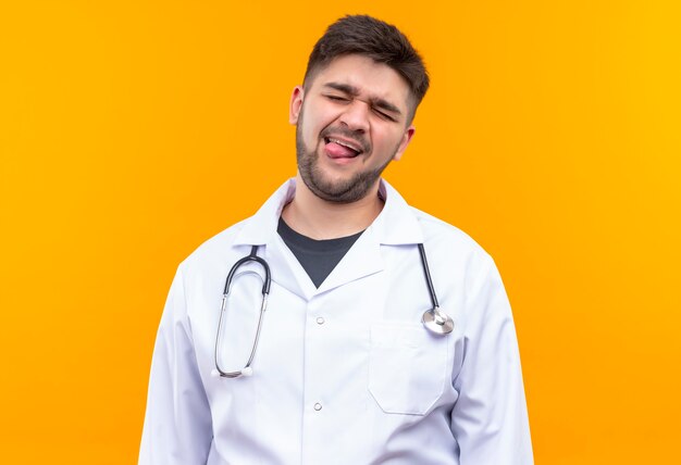 Joven médico guapo con bata médica blanca, guantes médicos blancos y un estetoscopio cerrando los ojos juguetonamente mostrando el ratón parado sobre la pared naranja