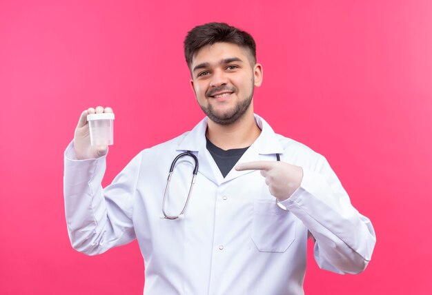 Joven médico guapo con bata médica blanca, guantes médicos blancos y un estetoscopio contento de los resultados que señalan en el contenedor de análisis transparente de pie sobre una pared rosa